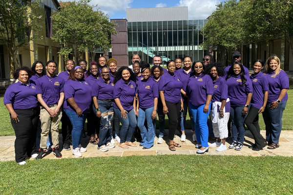 Large group of people all wearing purple polos. They are standing outside in front of a building. 