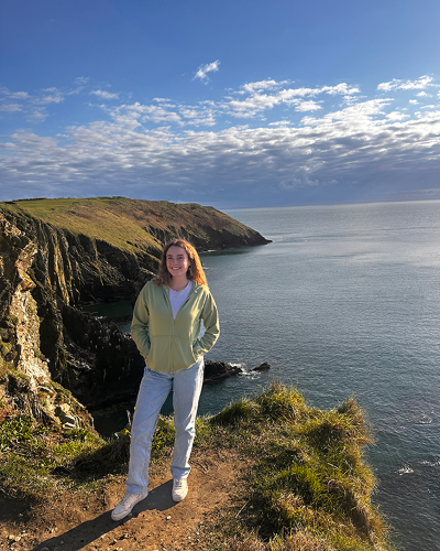 Kennedy Crouse stands by an oceanside cliff in Spain. She is dressed casually and smiles at the camera on a beautiful day.