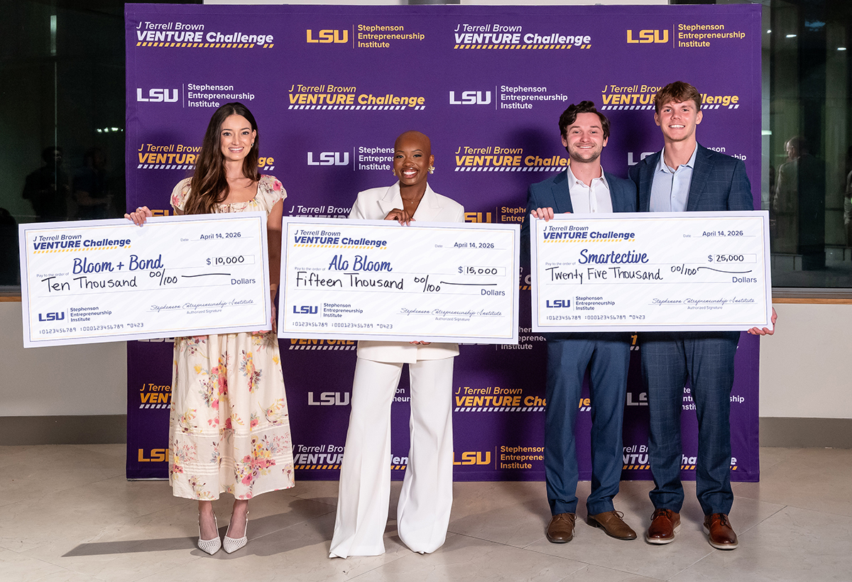 Winners of the J Terrell Brown Venture Challenge stand in front of a branded backdrop and hold large cardboard checks showing their winnings. All four are wearing business attire and smile warmly.
