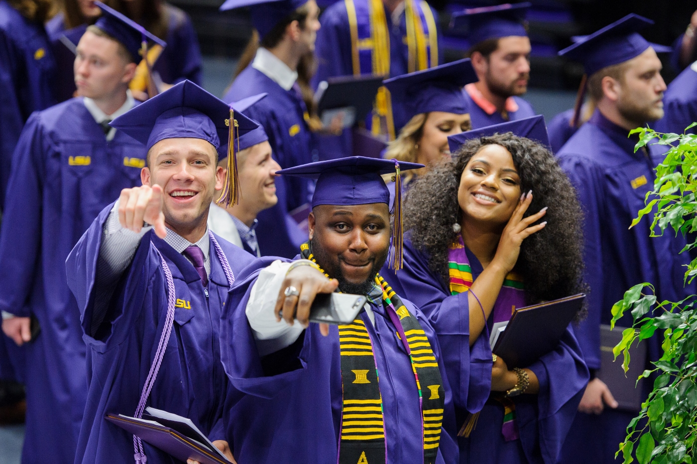 A group of graduates in purple gowns celebrate at a college commencement ceremony. They have joyous expressions, one individual points to the camera.